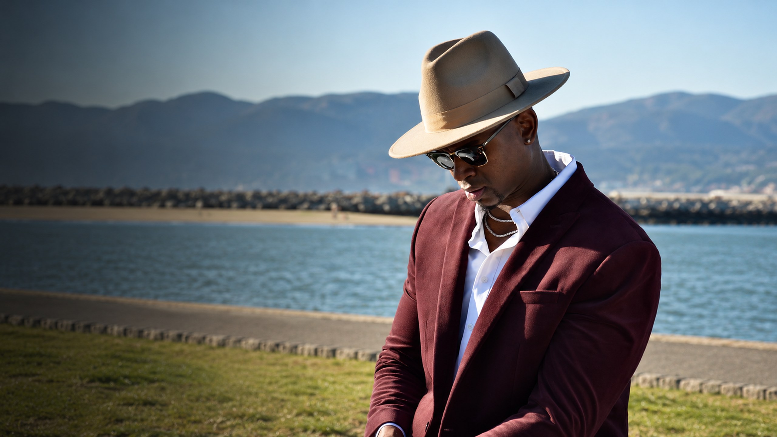 Michael K. Farley in a tan hat and burgundy blazer, looking down, with the bay and mountains behind him
